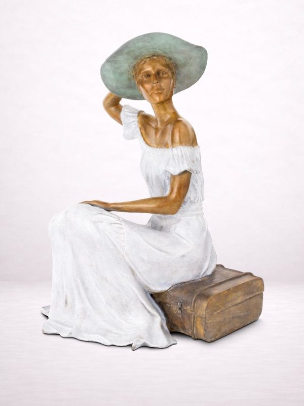 Sculpture en bronze d’une femme assise sur une valise, portant un chapeau, vue de trois-quarts.