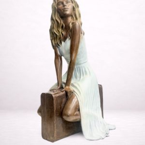 Sculpture en bronze d’une femme assise sur une valise, vue de trois-quarts.