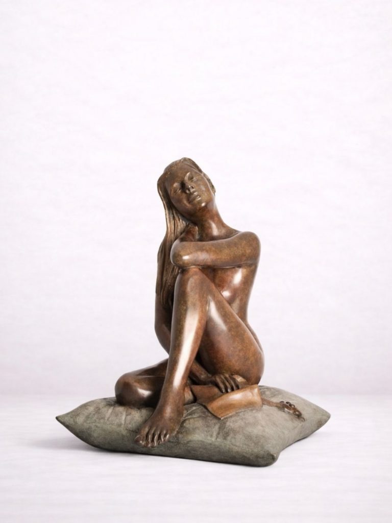 Sculpture en bronze d’une femme nue assise sur un coussin, une feuille à la main.