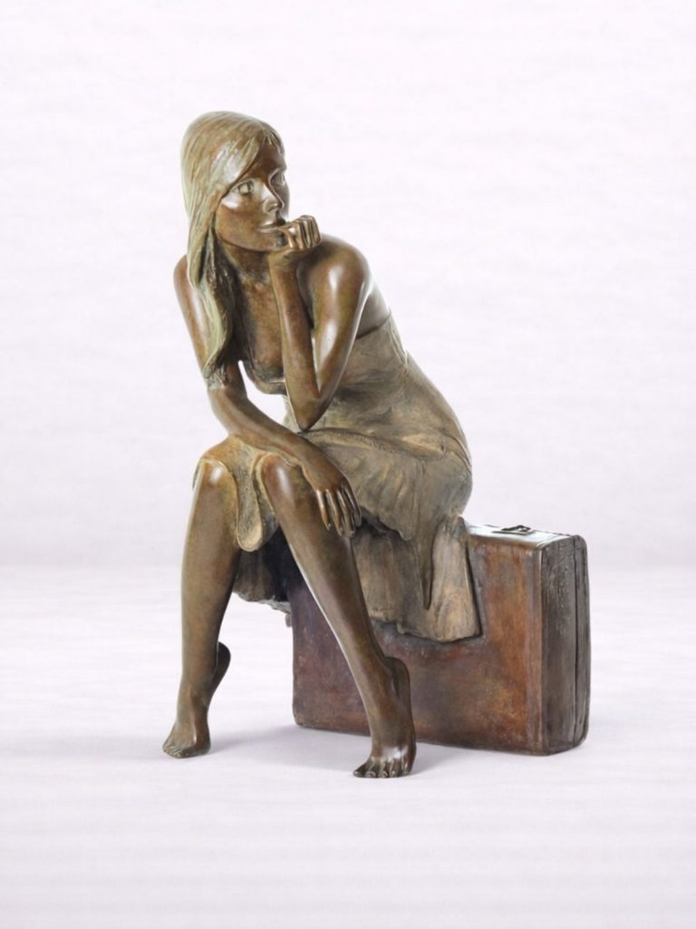 Sculpture en bronze d’une femme assise sur une valise, accoudée sur un genou, vue de face.