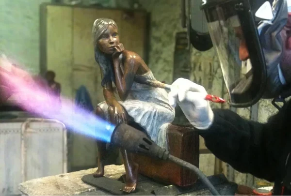 Application de patine sur une sculpture en bronze représentant une femme assise, avec une flamme de chalumeau et un pinceau.